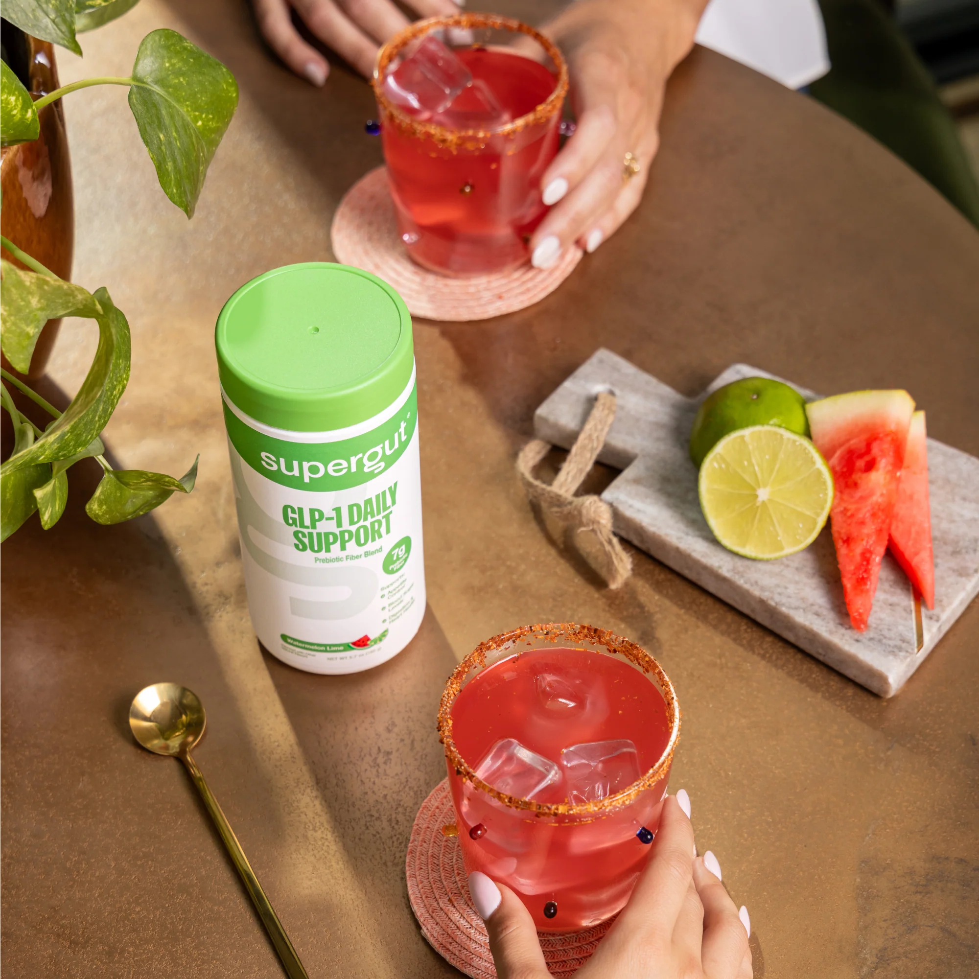 Two hands hold glasses of a red iced drink on a table with a Supergut GLP-1 Daily Support container, watermelon slices, lime, and a gold spoon.