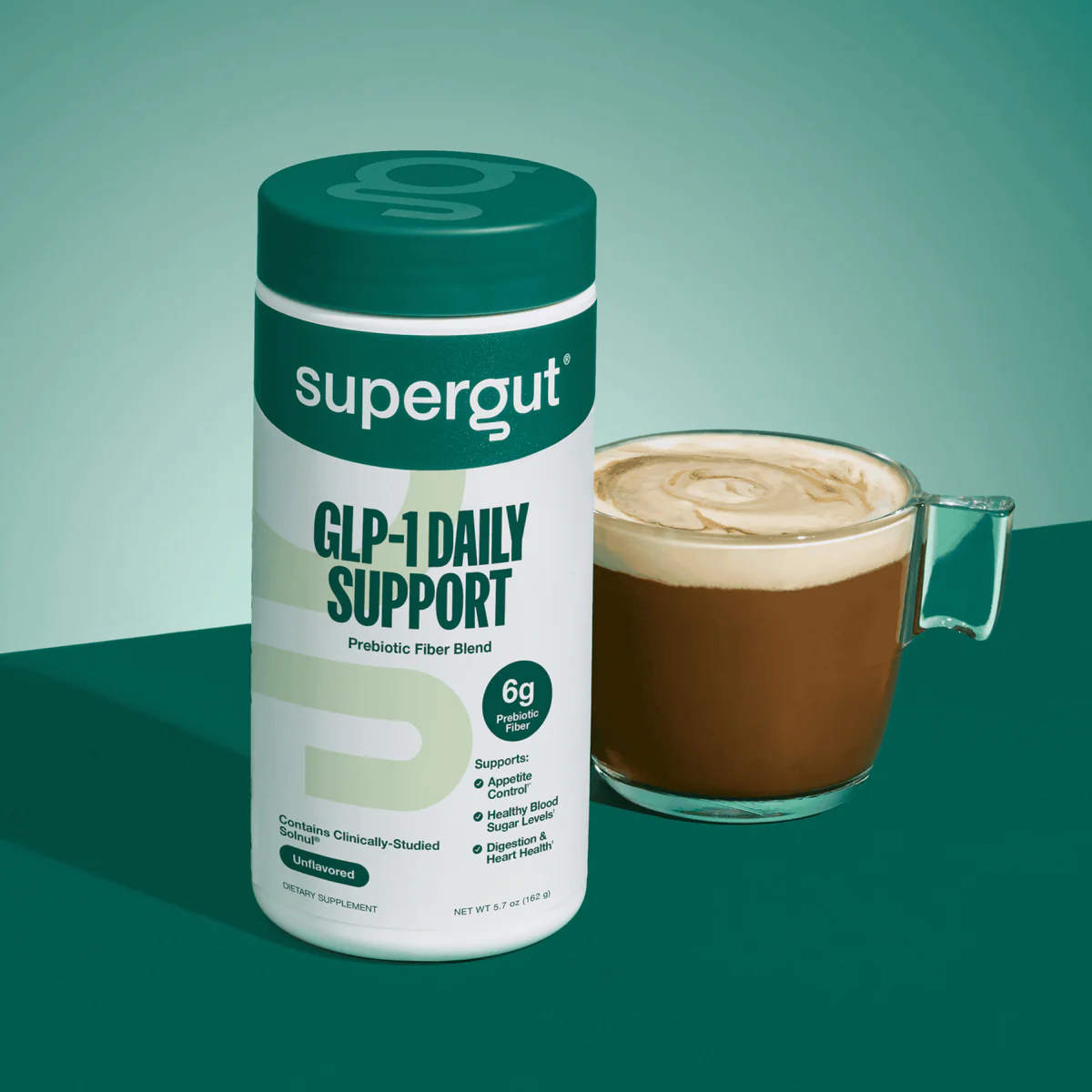 A container of Supergut GLP-1 Daily Support prebiotic fiber blend stands next to a glass mug of coffee with frothy milk on a green surface and background.
