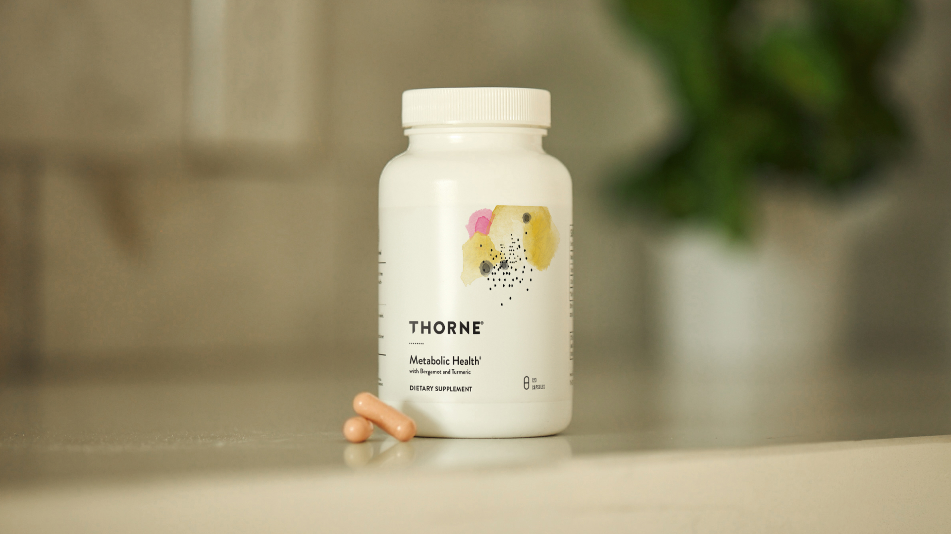 A white bottle labeled "Thorne Metabolic Health" sits on a surface with two beige capsules beside it; a blurred green plant is in the background.
