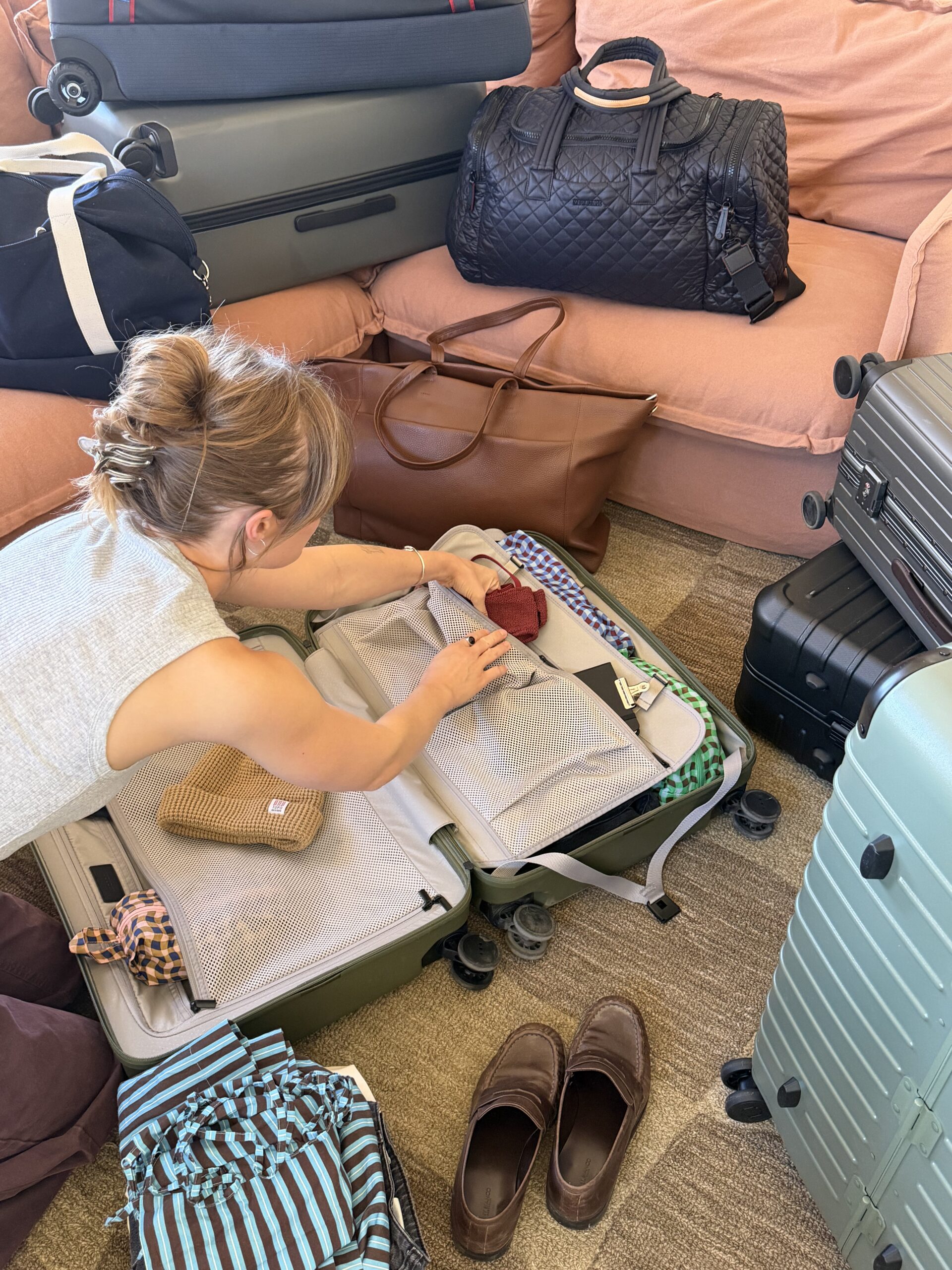 A person is packing clothes and small items into an open suitcase, surrounded by several bags and suitcases on a carpeted floor near a pink sofa.