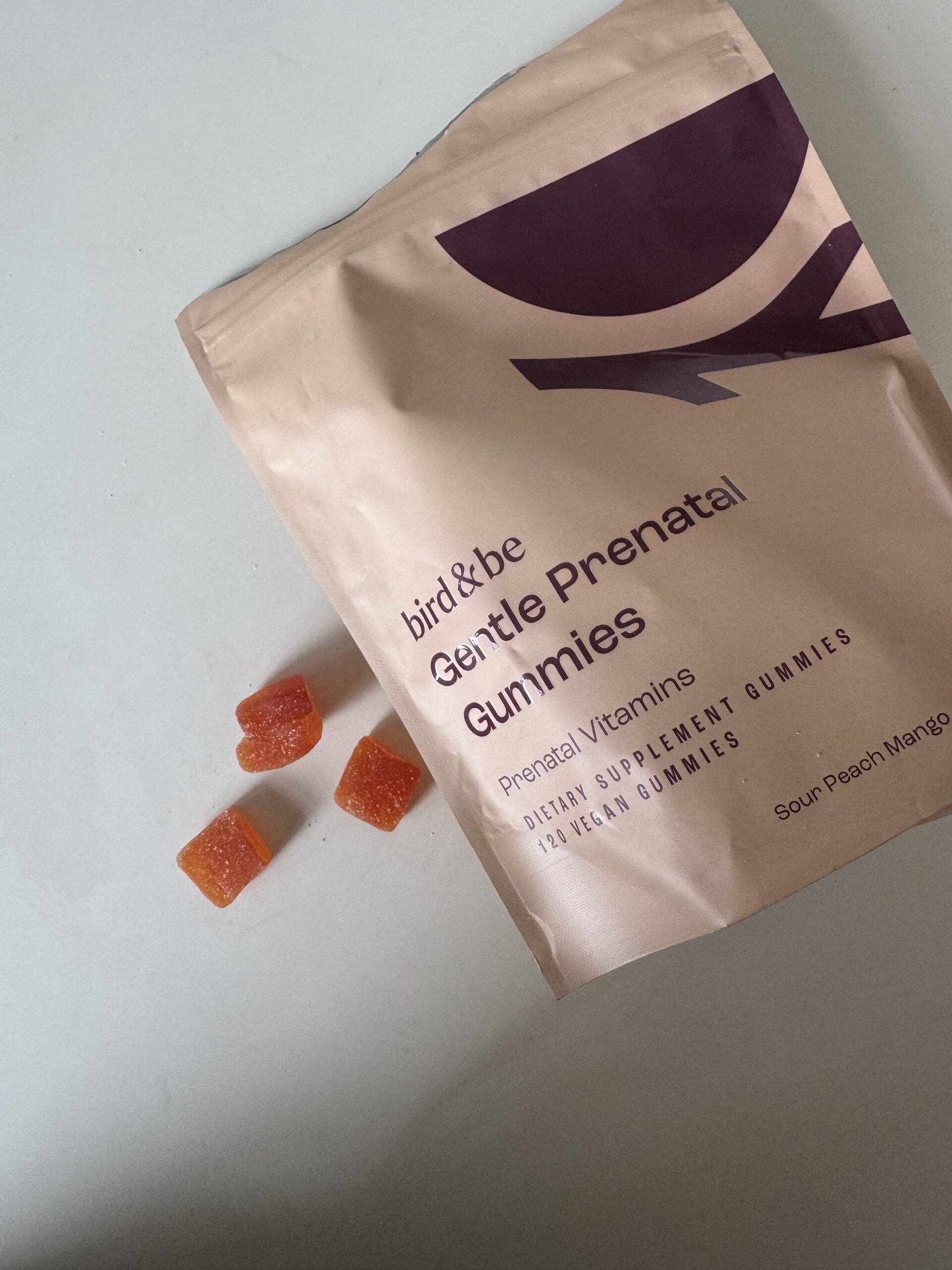 A beige pouch labeled “Gentle Prenatal Gummies” with four orange gummies scattered beside it on a white surface.