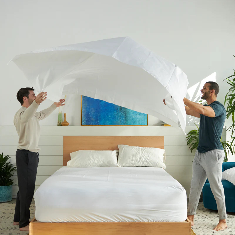 Two people are making a bed together, lifting a white cooling sheet over a neatly made bed in a bright, modern bedroom.