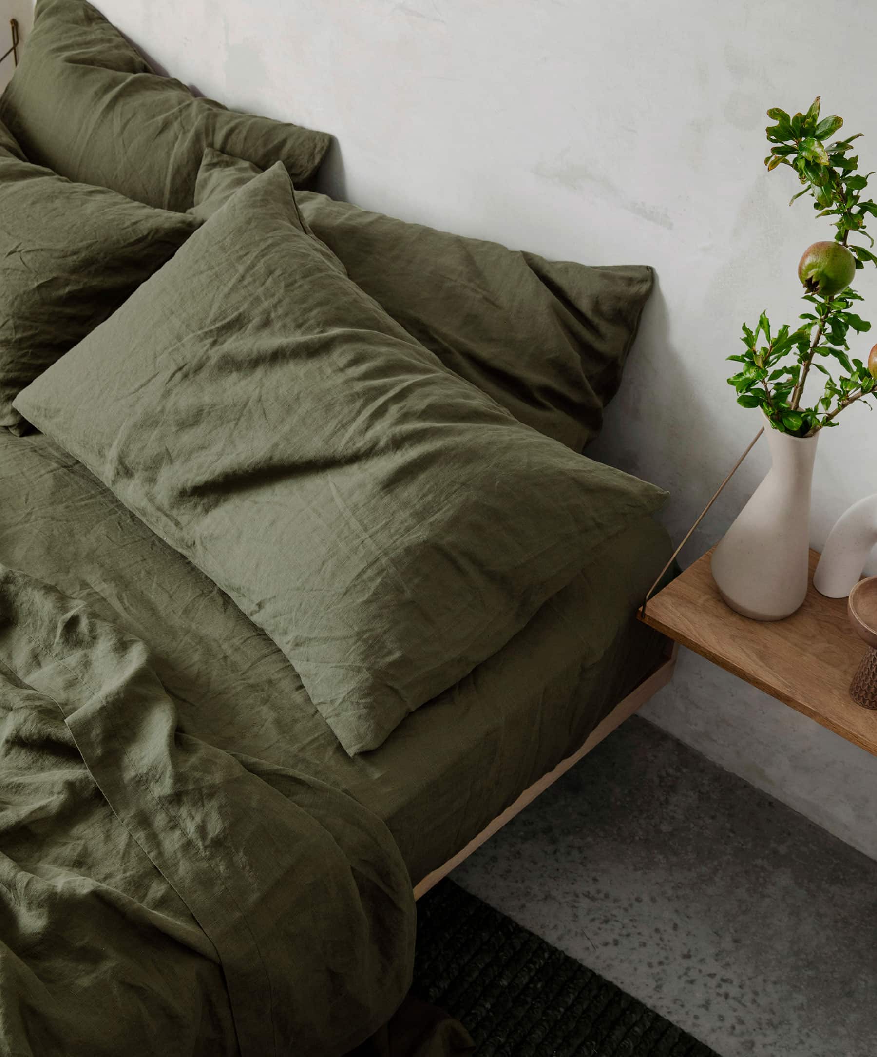 A bed with wrinkled olive green cooling sheets and pillows sits next to a wooden bedside table holding a white vase with green branches.