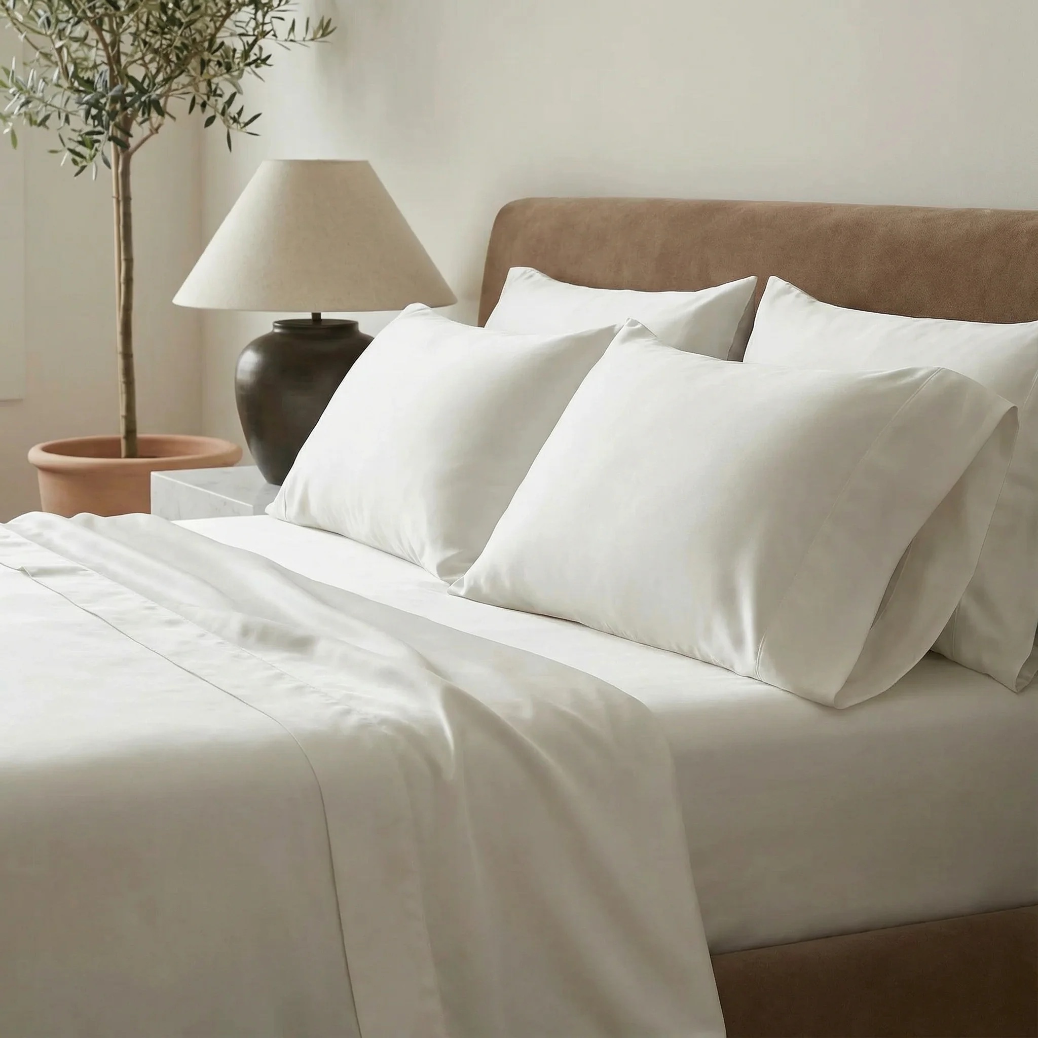 A neatly made bed with cooling sheets and pillows, a brown upholstered headboard, a bedside table with a lamp, and a potted plant in the background.