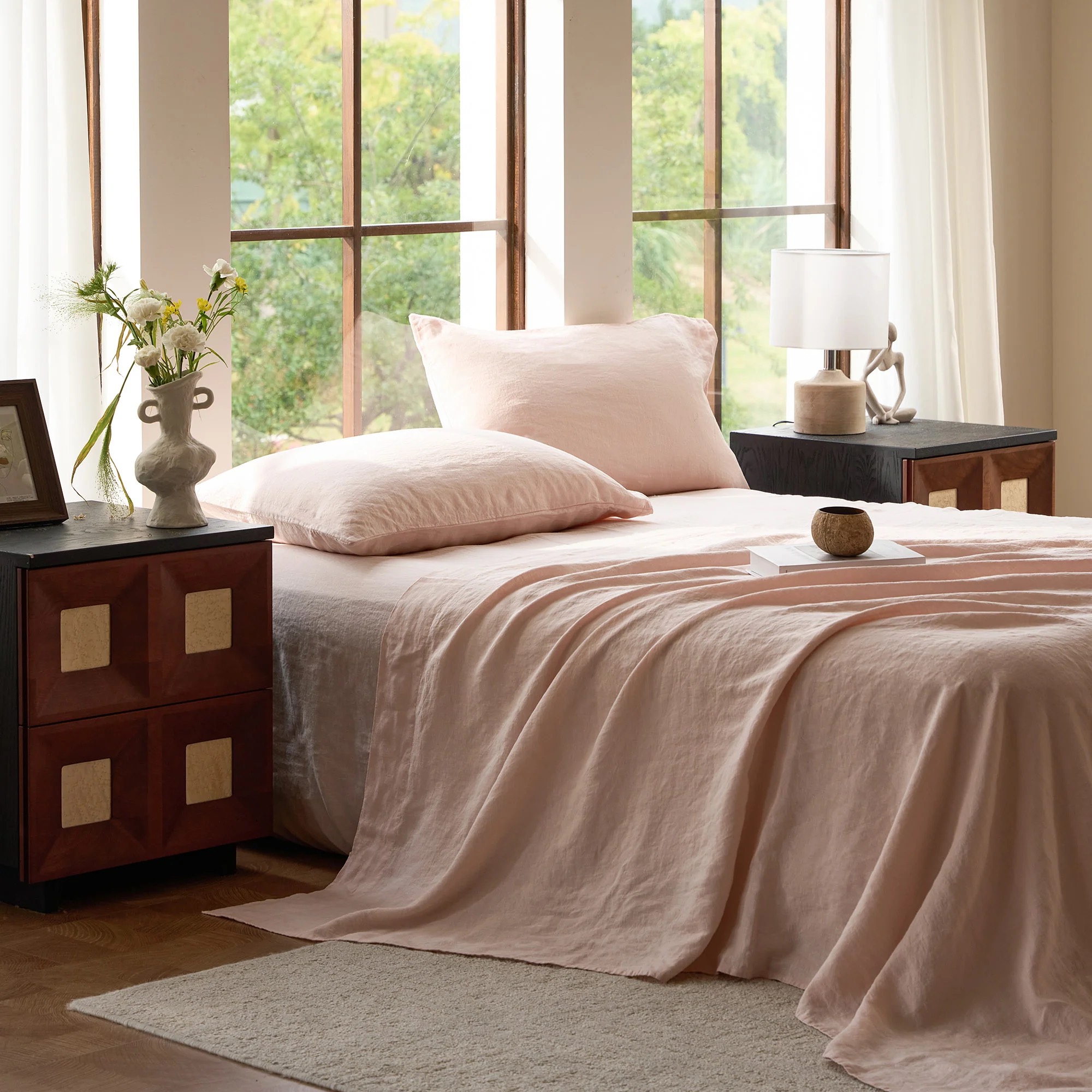 A neatly made bed with pale pink cooling sheets is positioned by large windows, flanked by dark wood nightstands with lamps and decorative items, in a bright, airy bedroom.