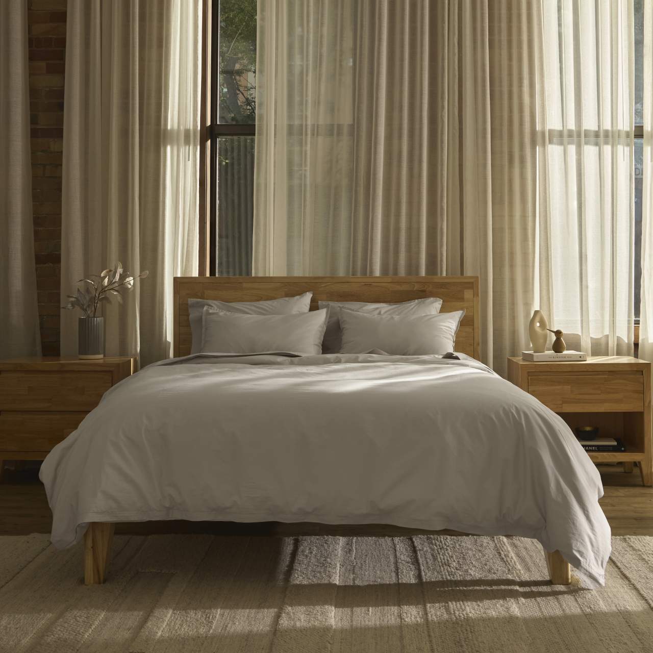 A neatly made bed with white cooling sheets is centered between two wooden nightstands in a sunlit bedroom with sheer curtains and large windows.