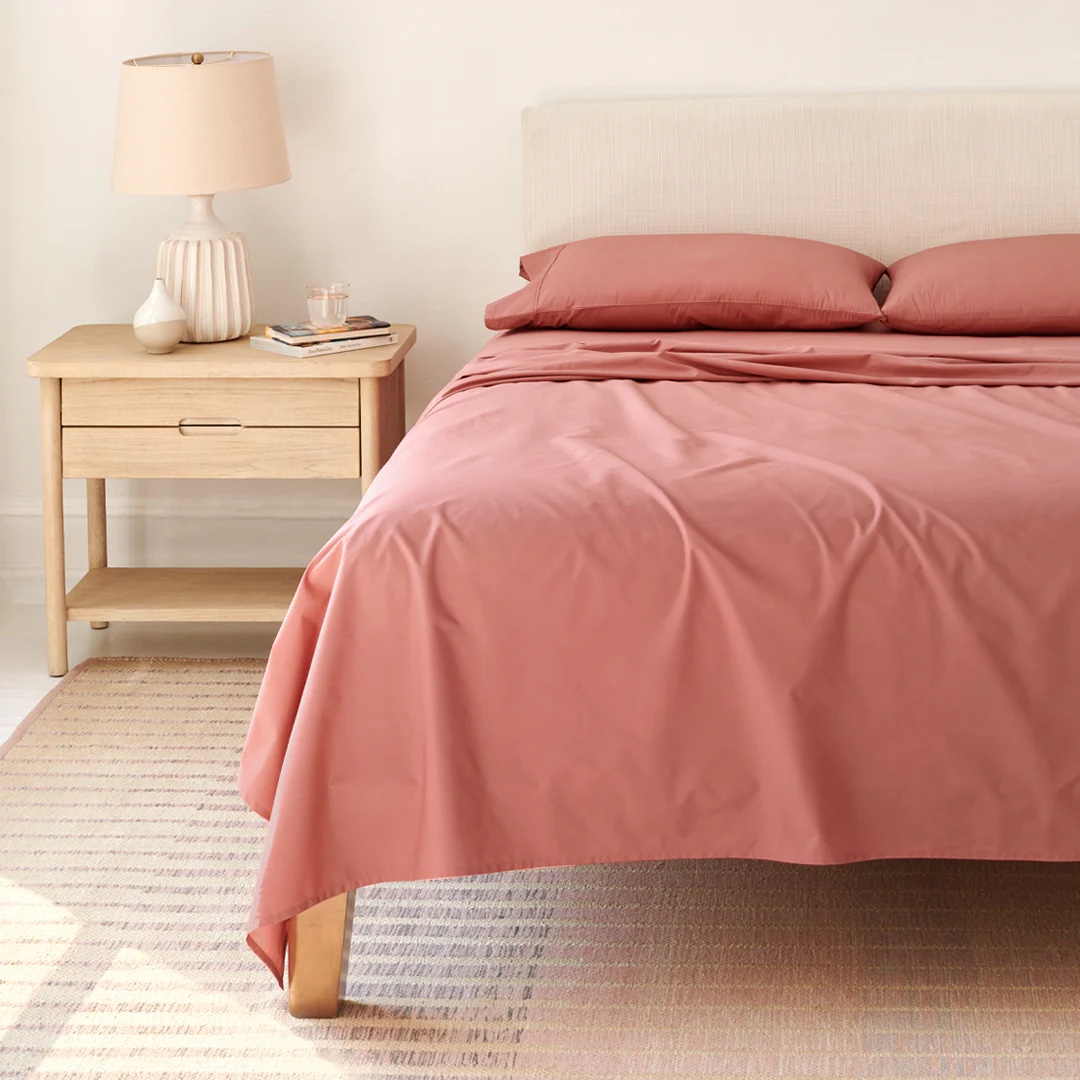 A bed with rose-colored cooling sheets and pillows sits next to a wooden nightstand holding a lamp, book, and cup, all set atop a light rug.