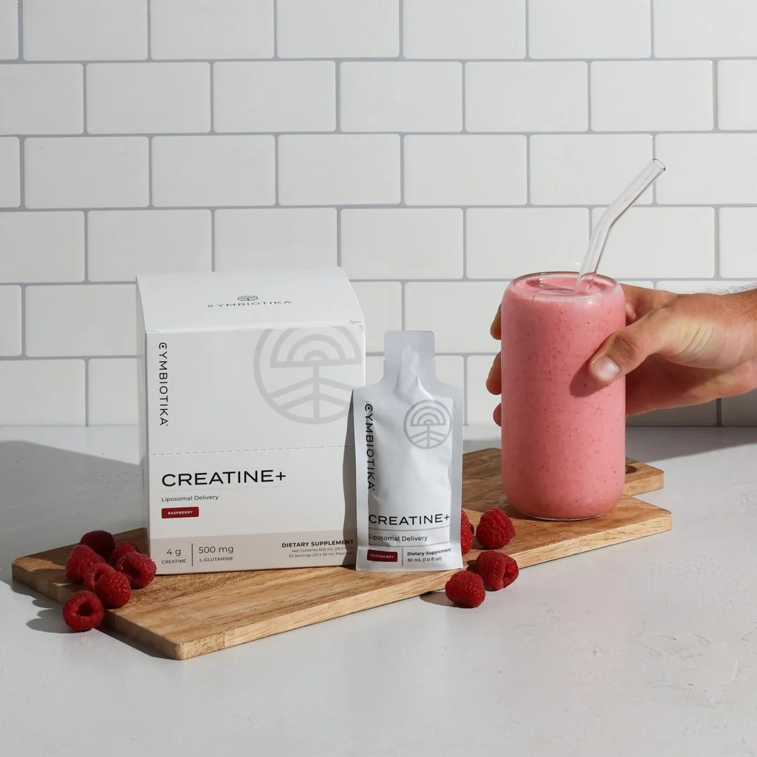 A hand holds a glass of pink smoothie with a glass straw next to a box and sachet of Cymbiotika Creatine+, raspberries, and a wooden board on a countertop.