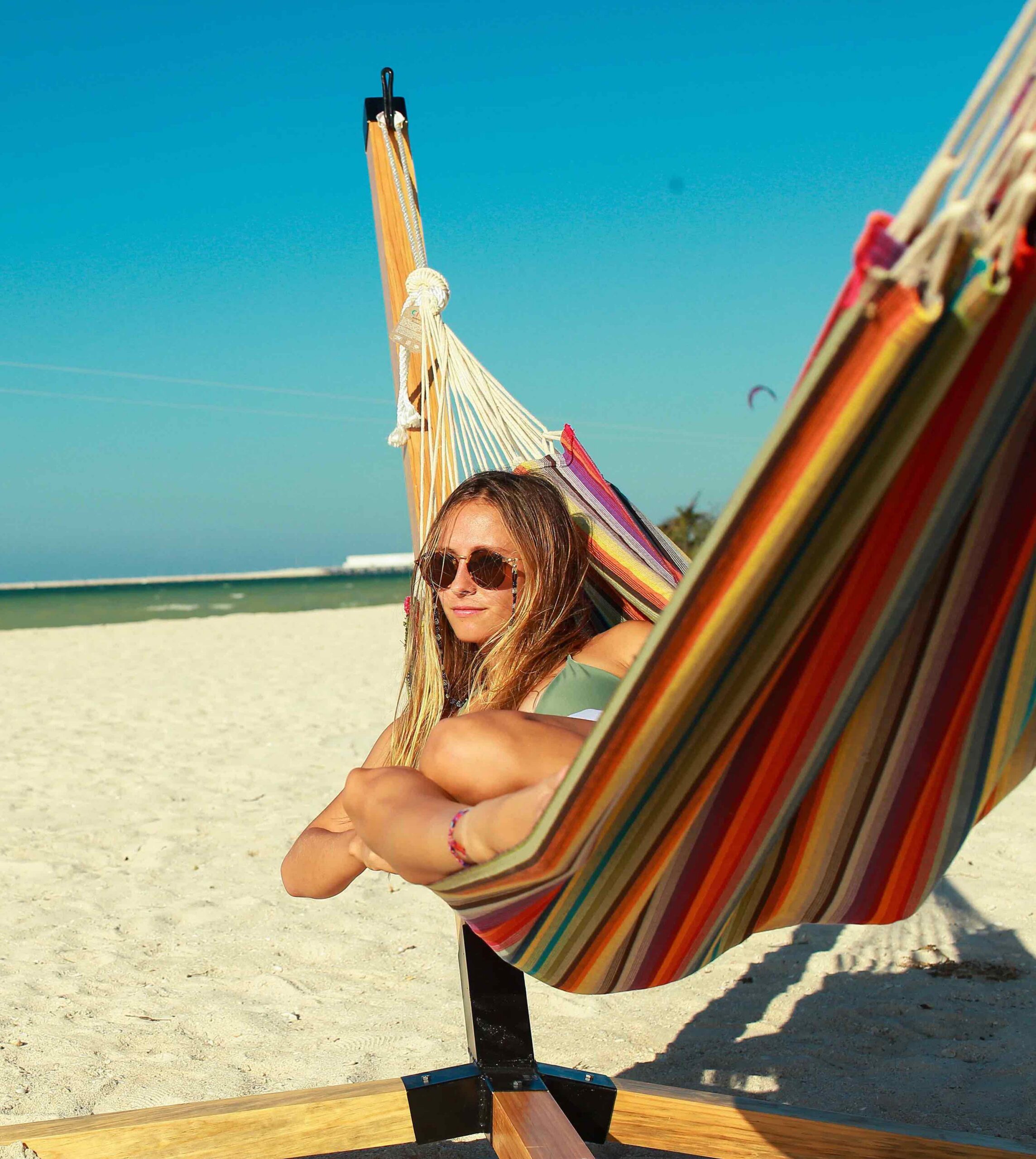 A person wearing sunglasses sits in a colorful hammock on a sandy beach with clear blue sky and ocean in the background.