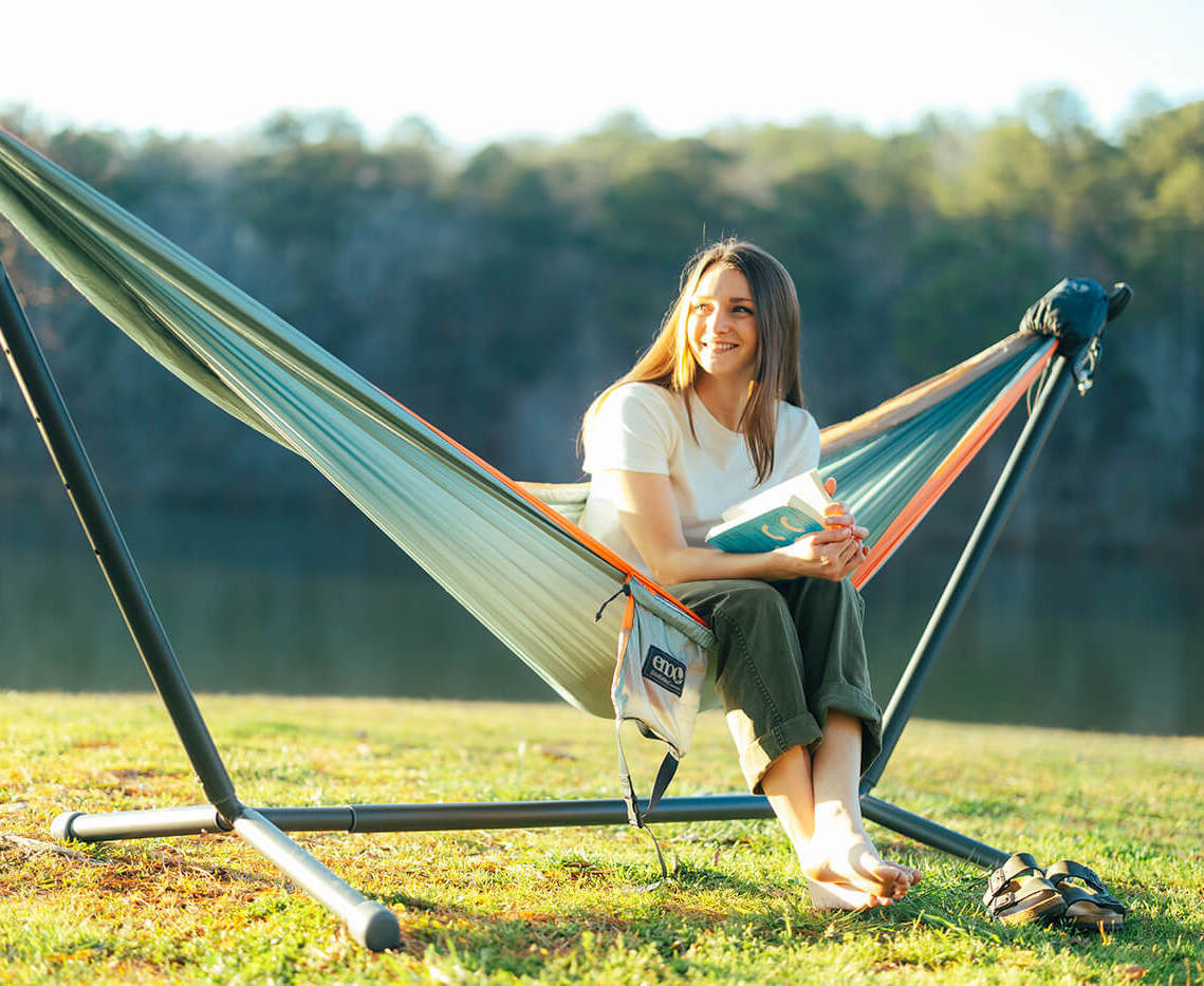 A woman sits in a hammock on a stand, holding a book and smiling, with a lake and trees in the background on a sunny day.