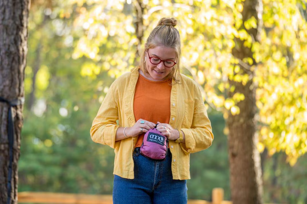 A woman in a yellow shirt and orange top stands outdoors, looking down while holding and opening a small pink pouch with both hands. Trees with yellow leaves are visible in the background.