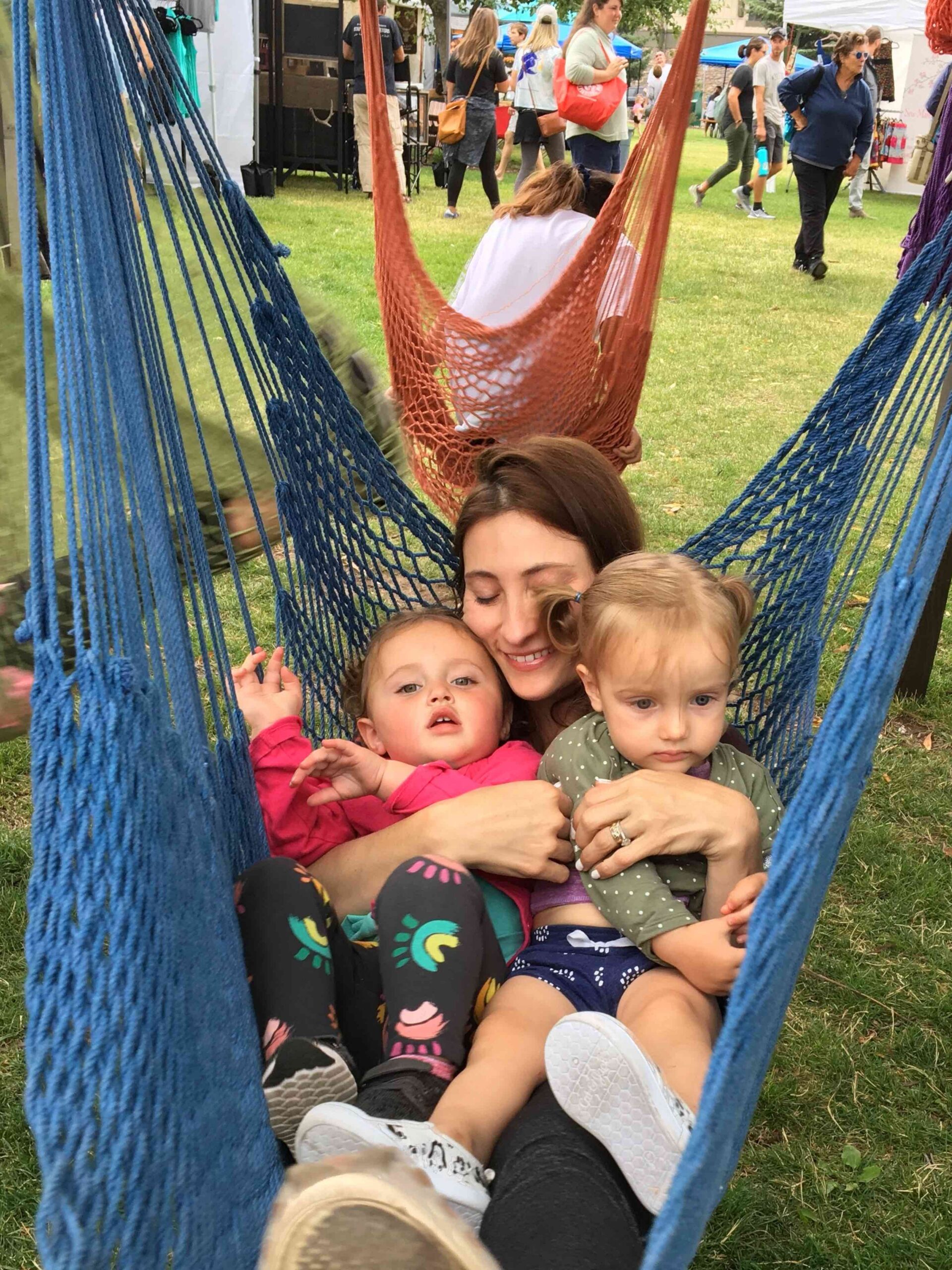 A woman sits in a blue hammock outdoors, holding two young children on her lap. Other people and hammocks are visible in the background.