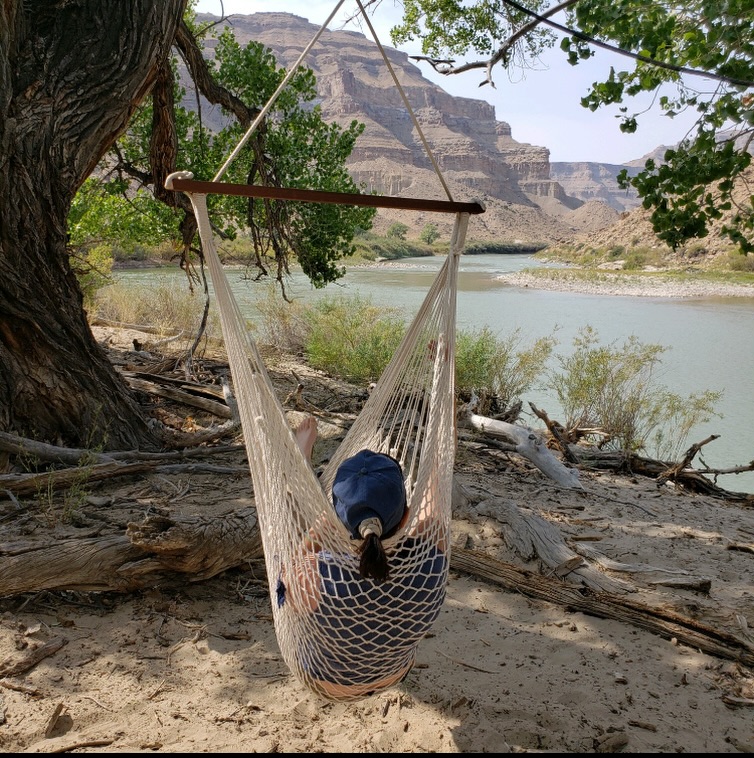 Person wearing a blue cap relaxes in a net hammock by a riverbank, surrounded by trees and rocky cliffs under a clear sky.