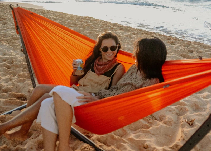 Two women sit in an orange hammock on a sandy beach, one holding a drink. They are facing each other and smiling, with the ocean visible in the background.