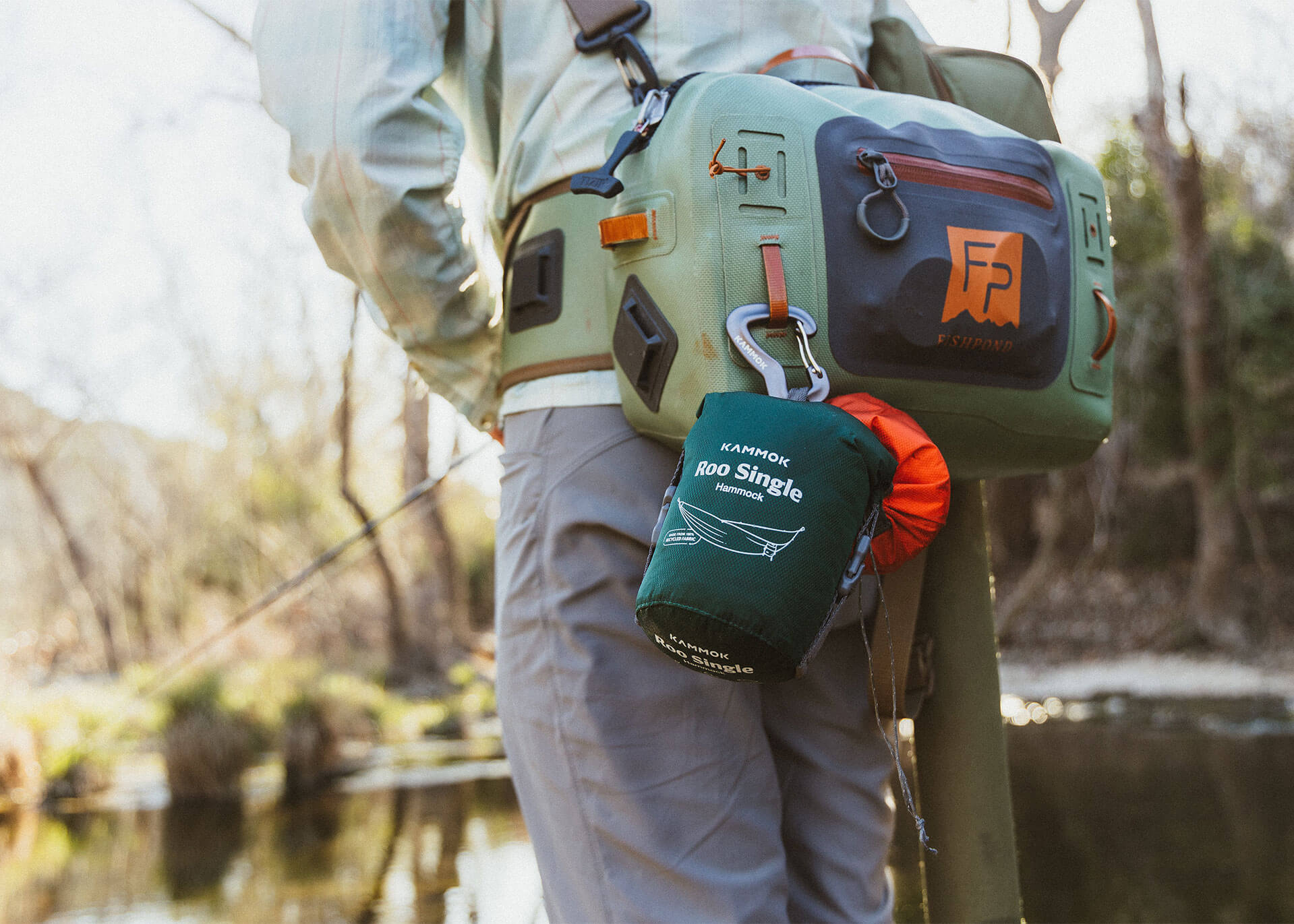 Person standing in shallow water wearing outdoor gear, with a fishing bag and a hammock pouch attached to their belt. Trees and water are visible in the background.