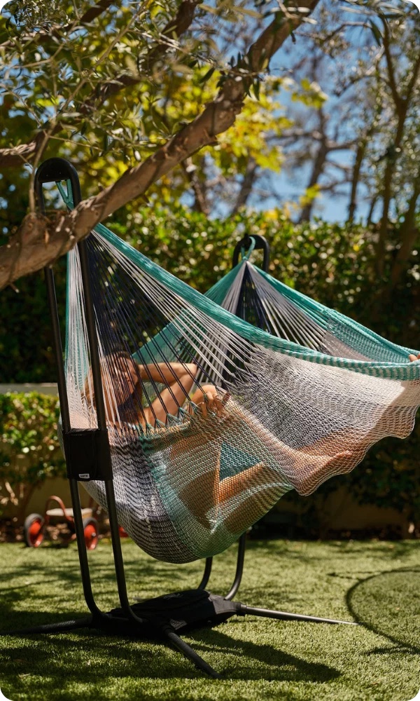 Person relaxing on a hammock in a sunlit garden with green grass, leafy trees, and blurred background foliage.