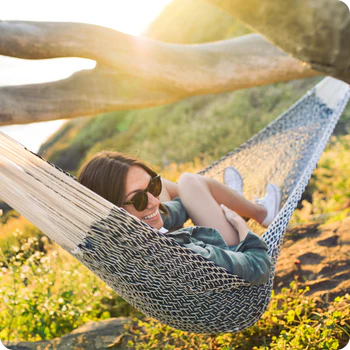 Woman wearing sunglasses lying on a hammock outdoors, surrounded by sunlight and nature, smiling and relaxing.