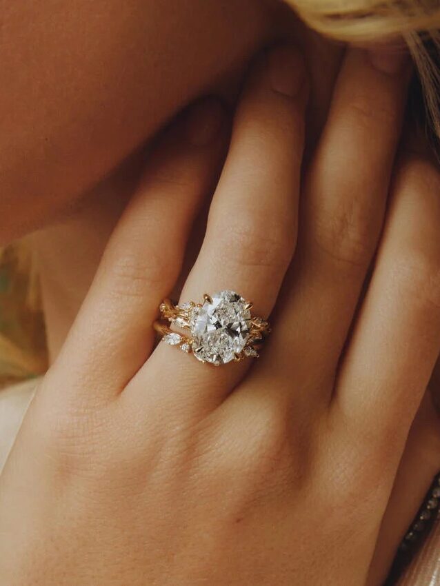 A close-up of a hand wearing two gold rings with a large oval diamond and smaller diamonds, set against the person's cheek.
