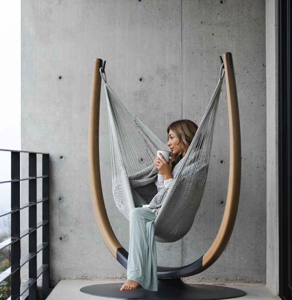 Woman sits in a hammock chair on a balcony, holding a cup and looking out, with a concrete wall and railing in the background.