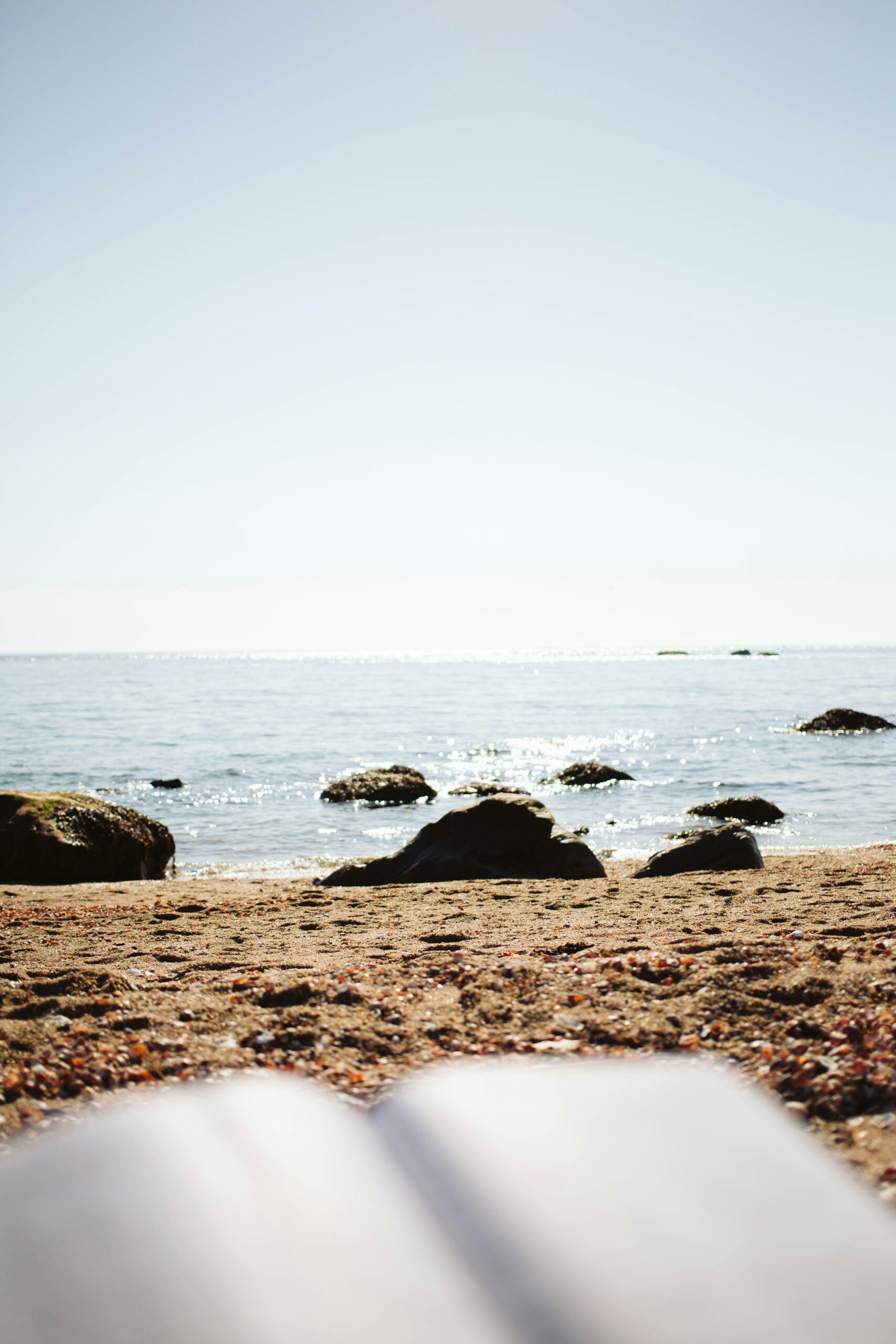 Rocky shoreline with small waves, sandy beach in the foreground, and a clear sky above.