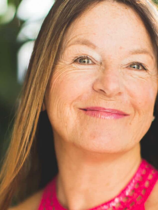 A woman with long straight hair and a pink top smiles at the camera, with green foliage and sunlight in the background.