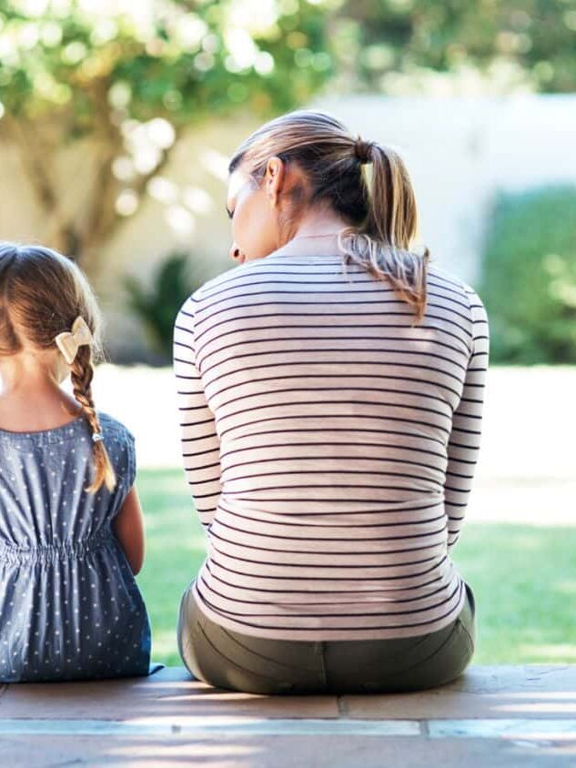 An adult and a young girl with braids sit on a porch facing a sunny backyard, both seen from behind.