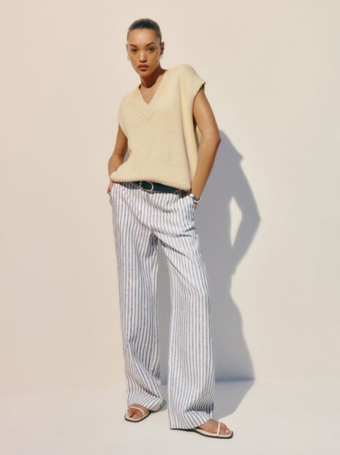 A woman stands against a plain background wearing a beige sleeveless top, loose striped pants, white sandals, and a black belt, with her hands in her pockets.