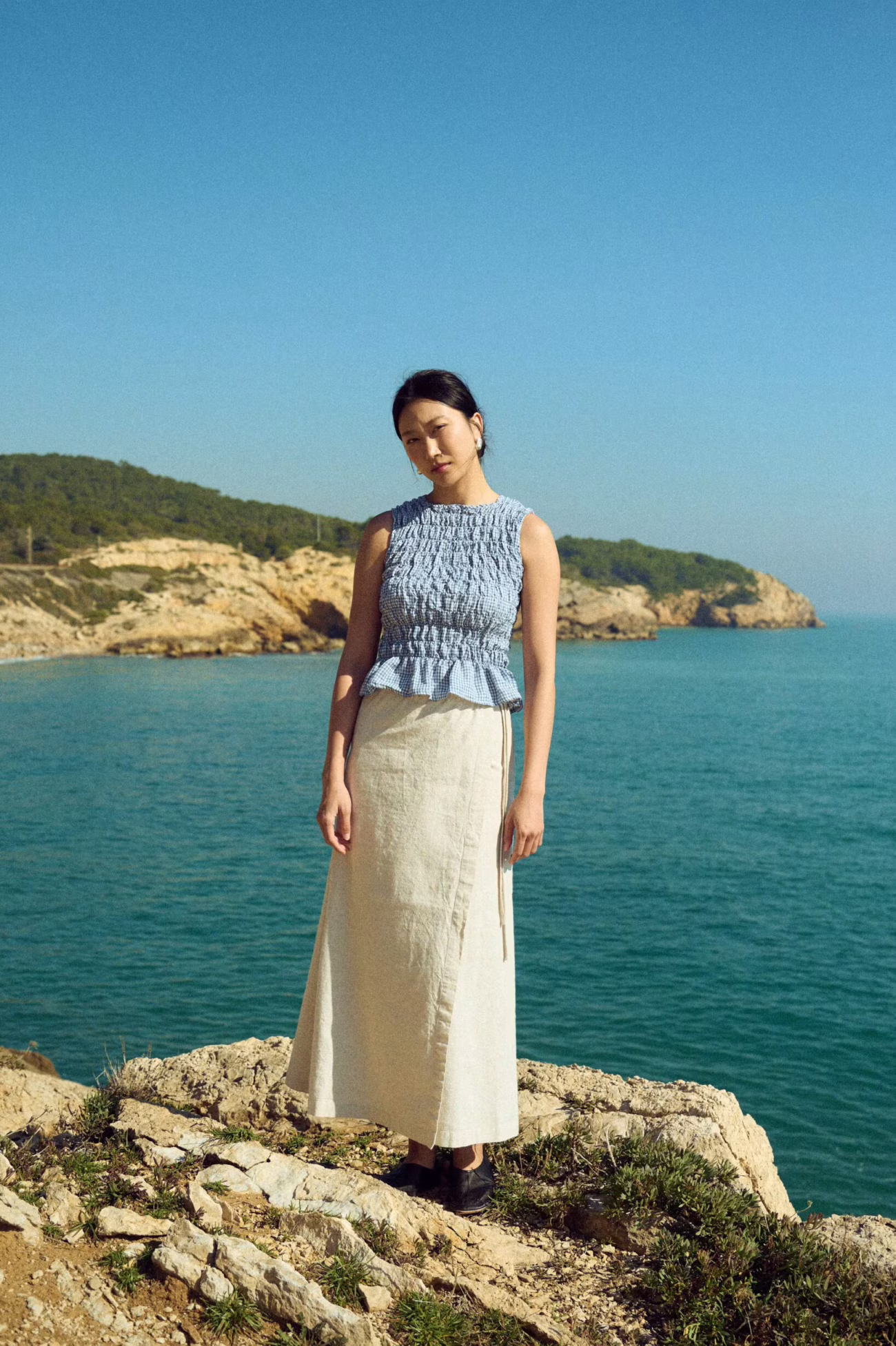 A woman stands on a rocky coastline wearing a light blue sleeveless top and a long beige skirt, with the sea and cliffs in the background under a clear sky.
