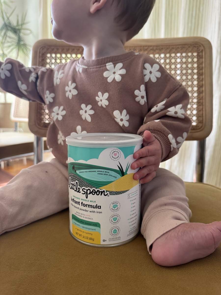 A young child sits on a chair holding a container of Little Spoon infant formula, wearing a brown floral sweater and light pants.