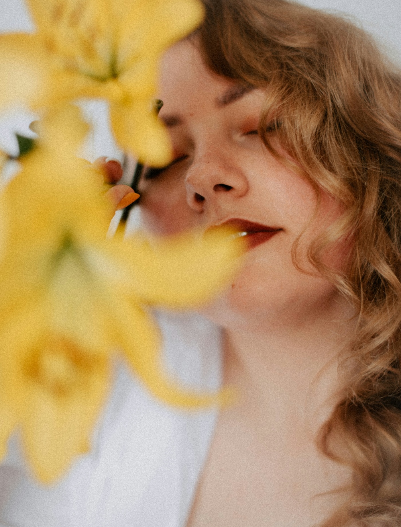 A person with wavy blonde hair and closed eyes holds a yellow flower close to their face, partially obscured by the petals in the foreground.