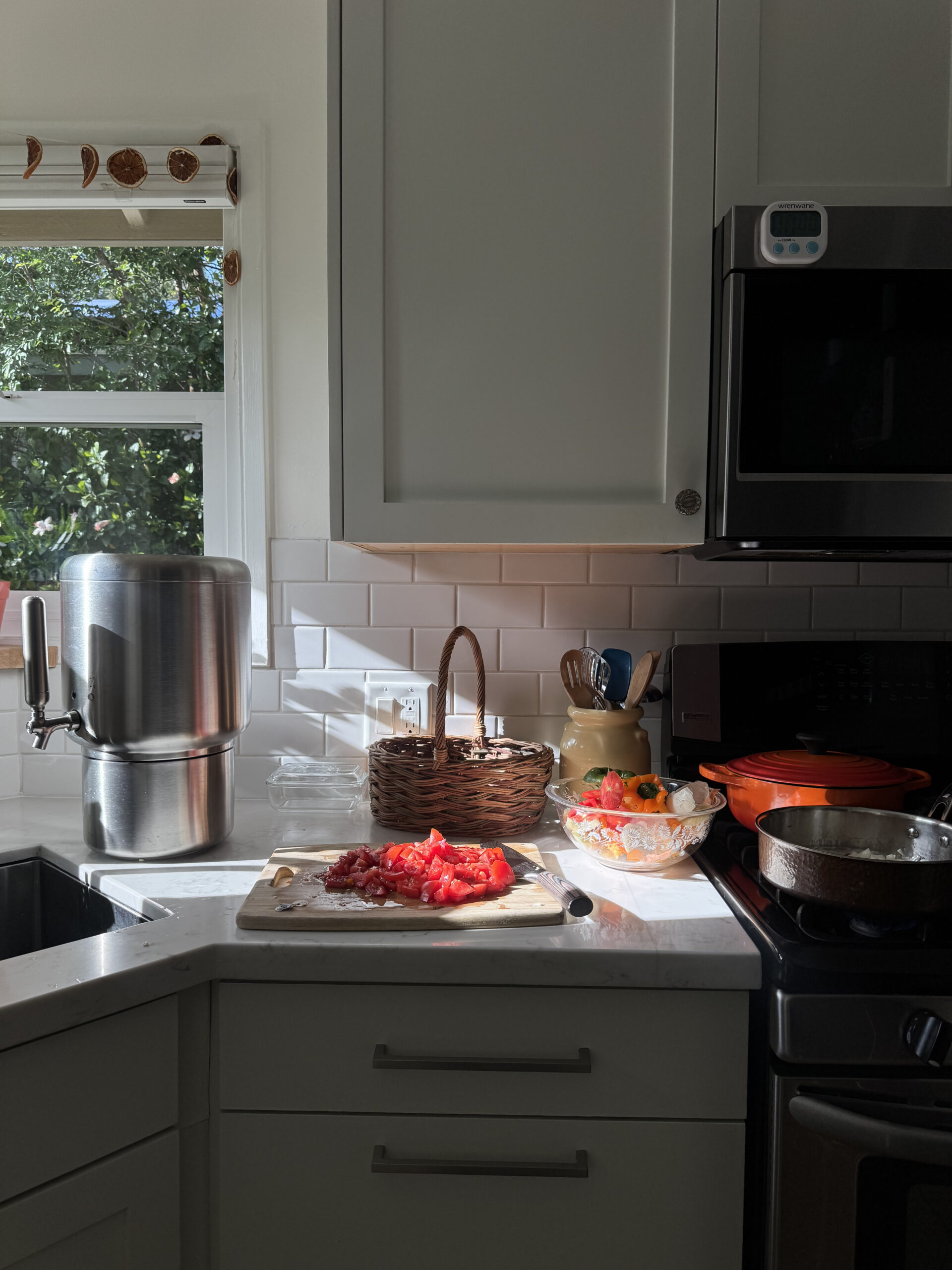 A kitchen countertop with chopped tomatoes on a cutting board, a bowl of vegetables, a pot on the stove, and a water filter near a sunny window.