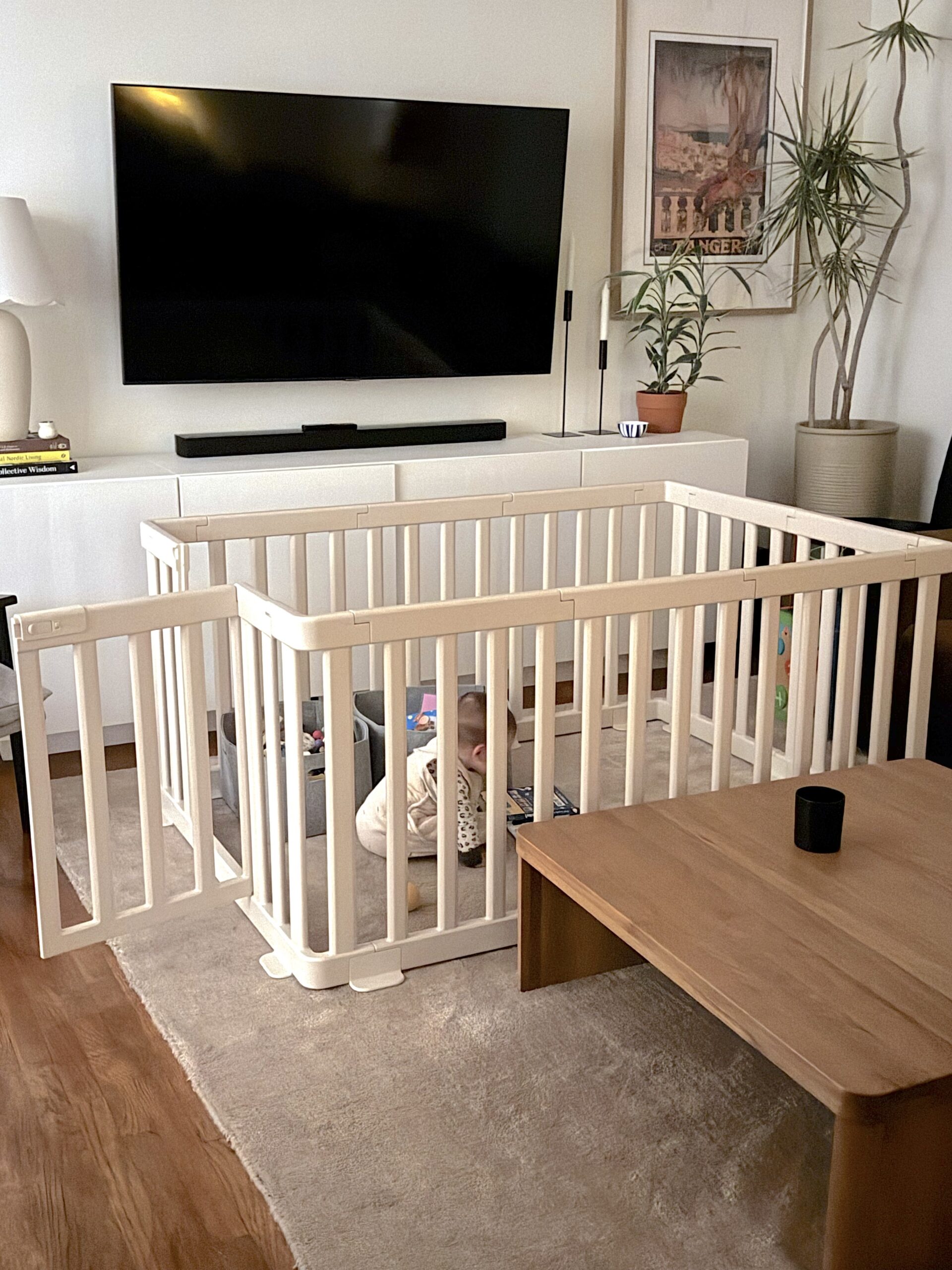 A baby sits and plays with toys inside a white safety playpen in a living room with modern furnishings and decor.