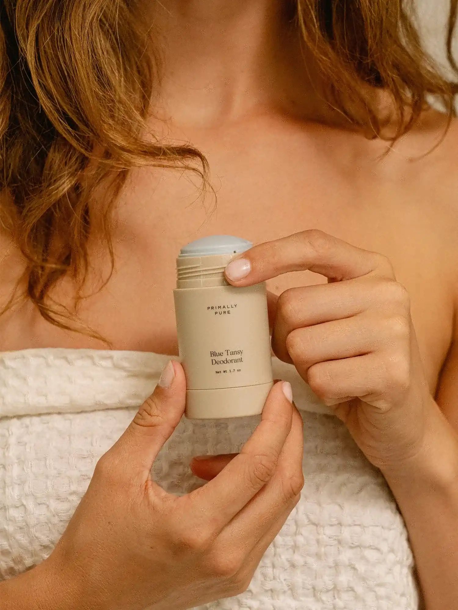 A person in a towel holds a stick of Blue Tansy Deodorant with one hand and is removing the cap with the other.