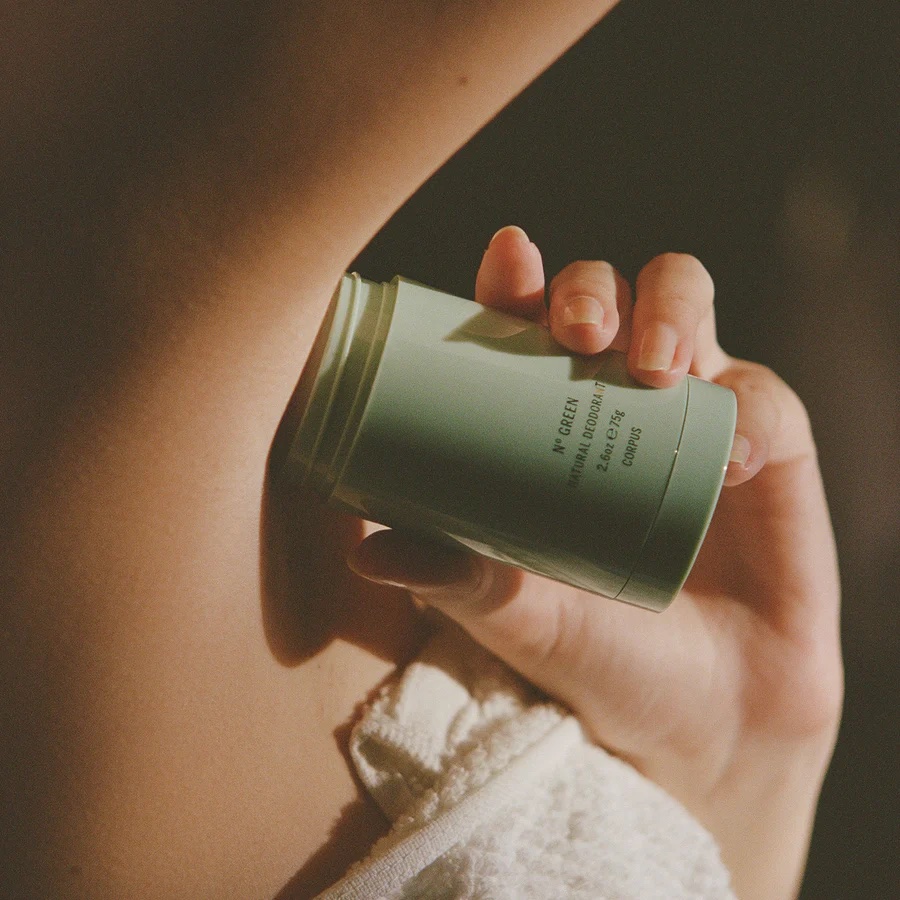 A person applies a green stick of deodorant to their underarm while wearing a white towel.