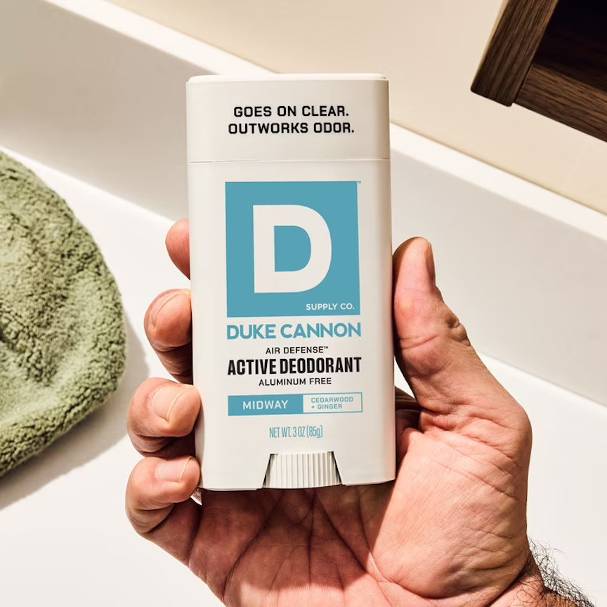 A hand holds a stick of Duke Cannon Active Deodorant in a bathroom, with a green towel and a white sink in the background.