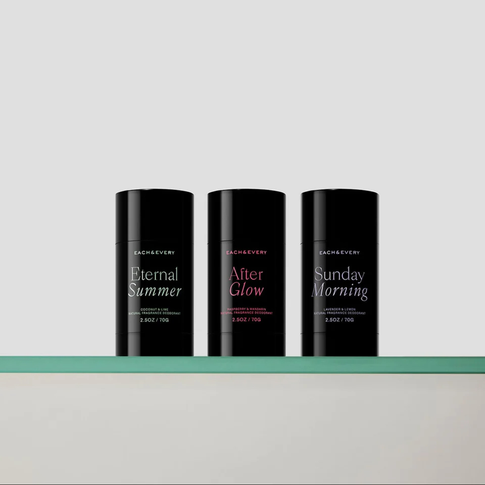 Three black cylindrical deodorant containers labeled Eternal Summer, After Glow, and Sunday Morning sit on a glass shelf against a plain light background.