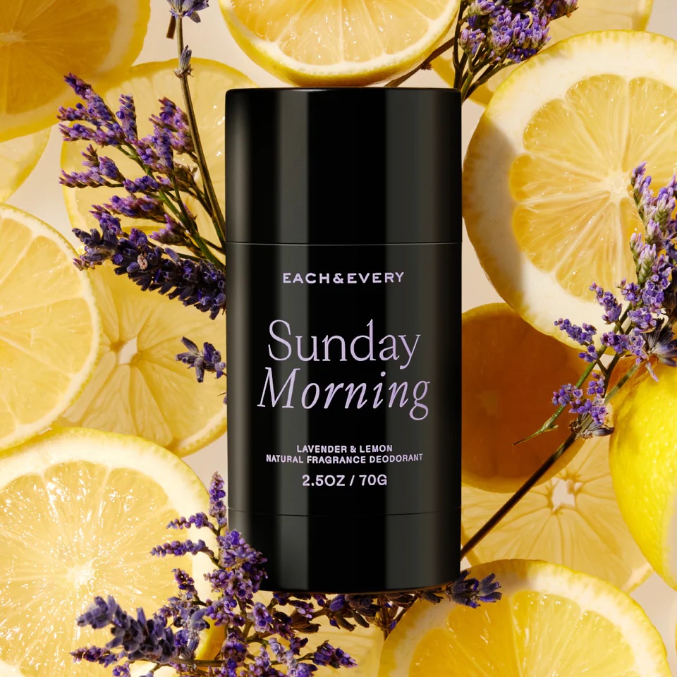 A black deodorant stick labeled "Sunday Morning" is surrounded by lemon slices and sprigs of lavender.