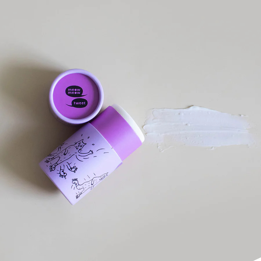 A purple container of Meow Meow Tweet deodorant with whimsical drawings, some product spread on a light surface, and the lid placed nearby.