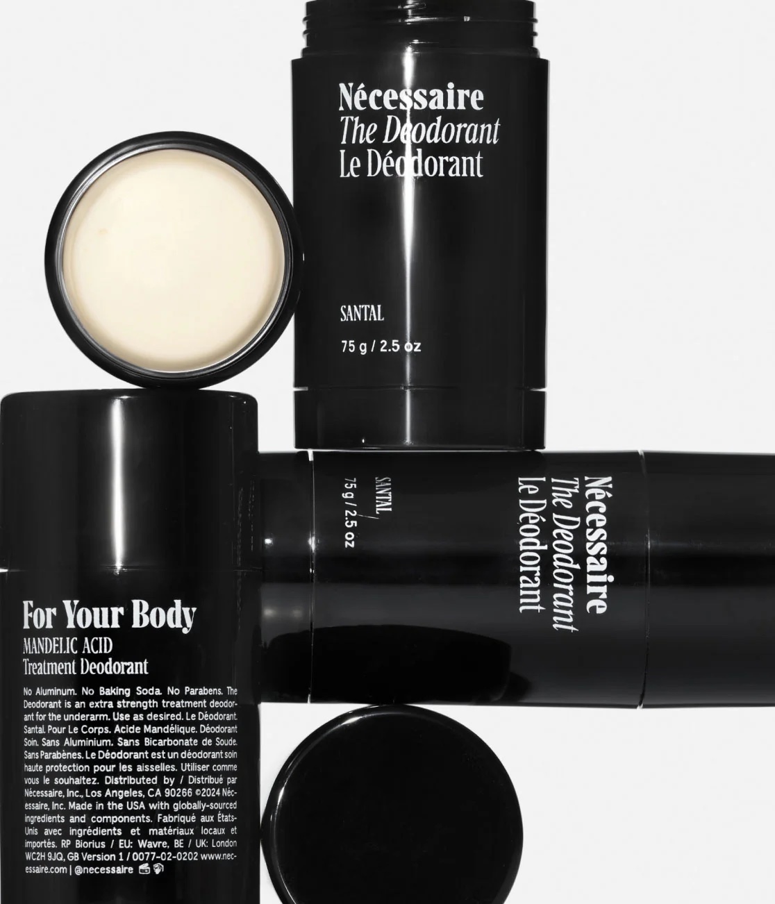Three black cylindrical containers of Nécessaire The Deodorant Santal are arranged on a white surface, one with its lid off showing the product inside. Labels display ingredients and product details.
