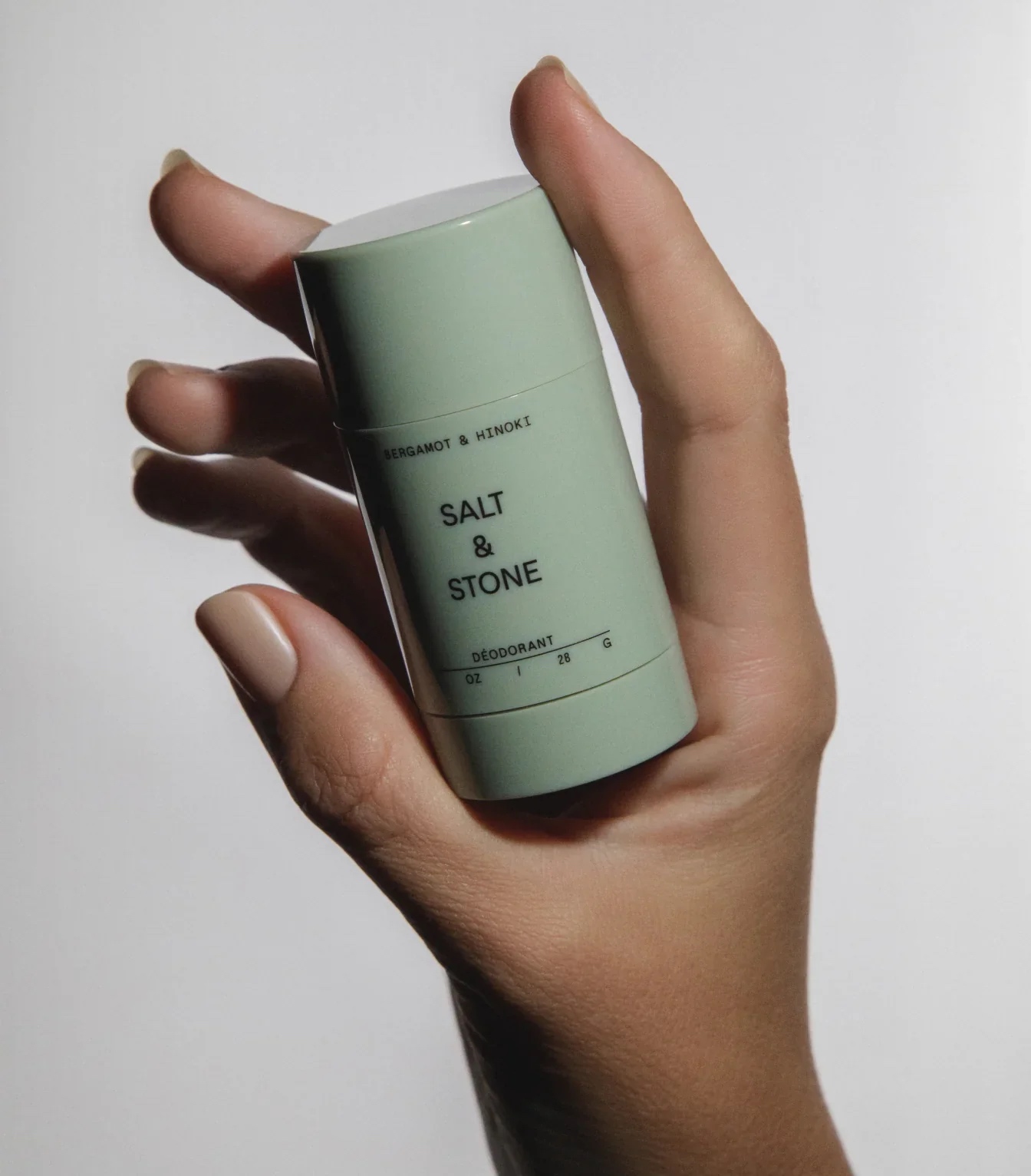 A hand holds a light green stick of Salt & Stone deodorant labeled "Bergamot & Hinoki" against a plain white background.