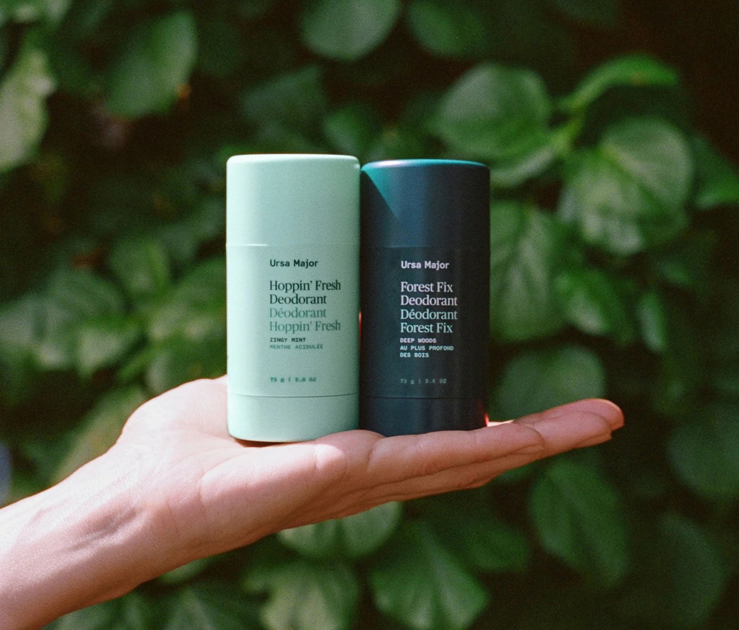 A hand holds two Ursa Major deodorant sticks, one light green labeled "Hoppin' Fresh" and one dark green labeled "Forest Fix," with leafy greenery in the background.