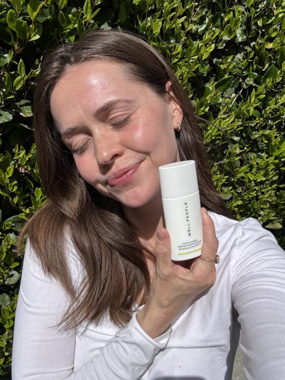A woman with long brown hair smiles with closed eyes in sunlight, holding a white bottle labeled "TRUE BOTANICALS" in front of green foliage.