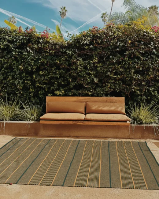 Outdoor scene with a tan cushioned bench against a leafy ivy wall, green plants, and a striped rug on the ground under a sunny sky.