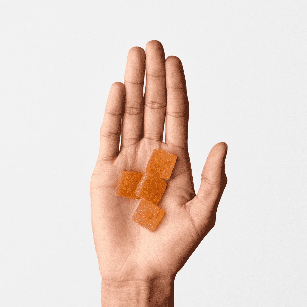 An open hand holding four square, orange, sugar-coated gummy candies against a plain white background.