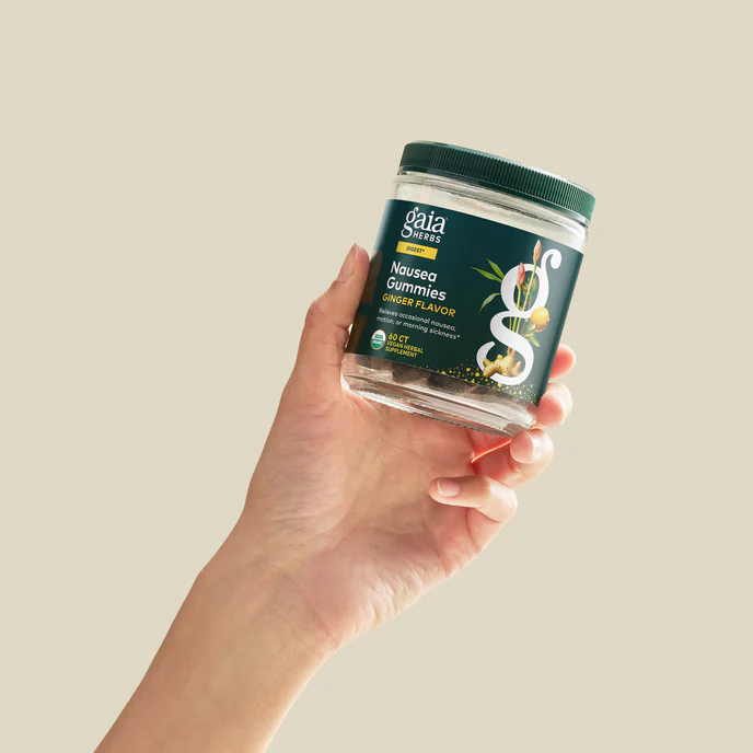 A hand holds a jar of Gaia Herbs Nausea Gummies, ginger flavor, against a plain beige background.