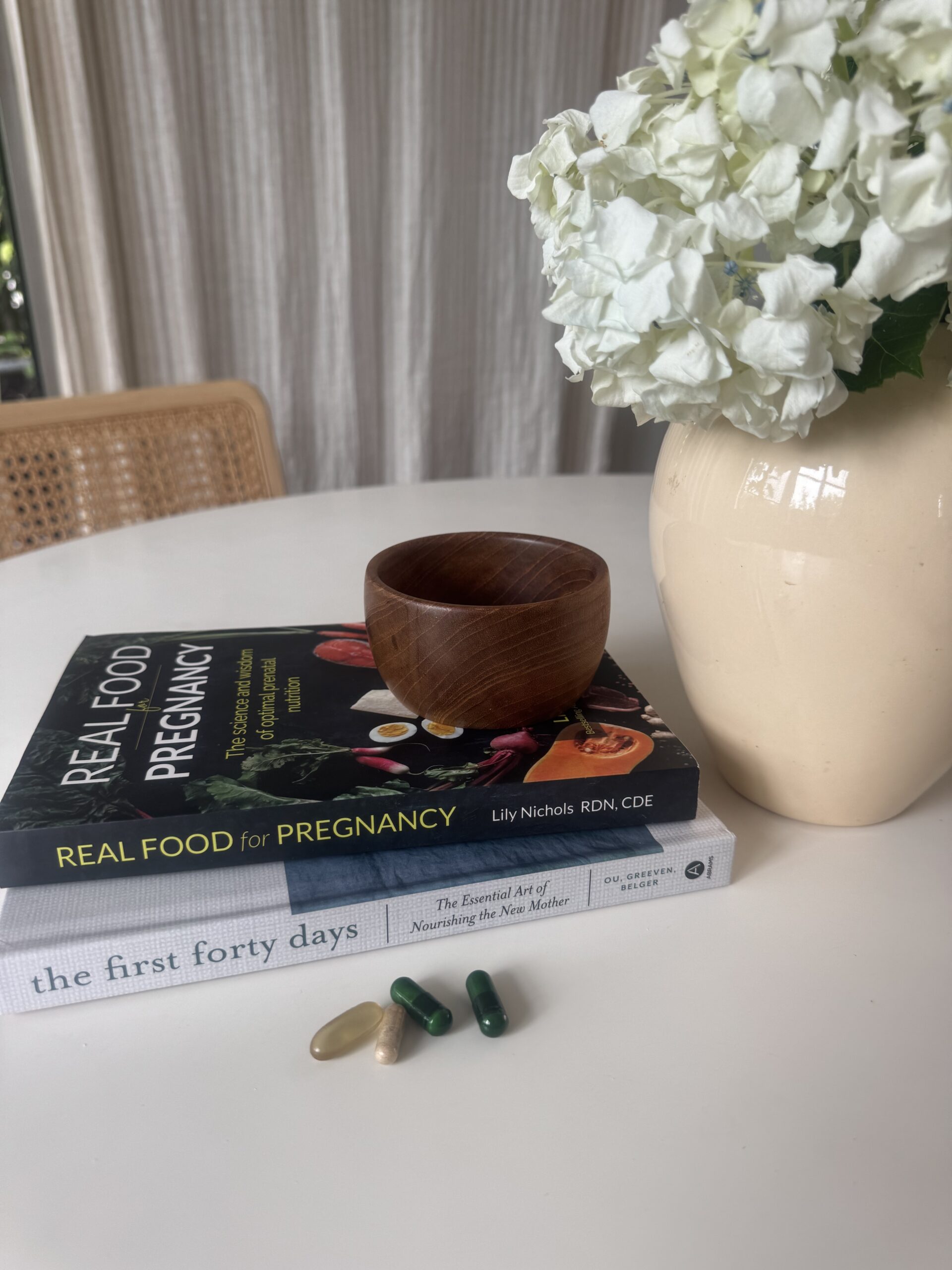 A stack of books, a wooden bowl, a vase of white flowers, and several prenatal vitamin capsules rest on a round white table.