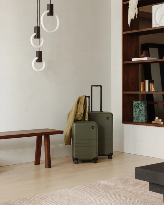 Two green suitcases and a khaki jacket are placed next to a wooden bench in a modern, minimalistic living space with shelving and contemporary lighting.
