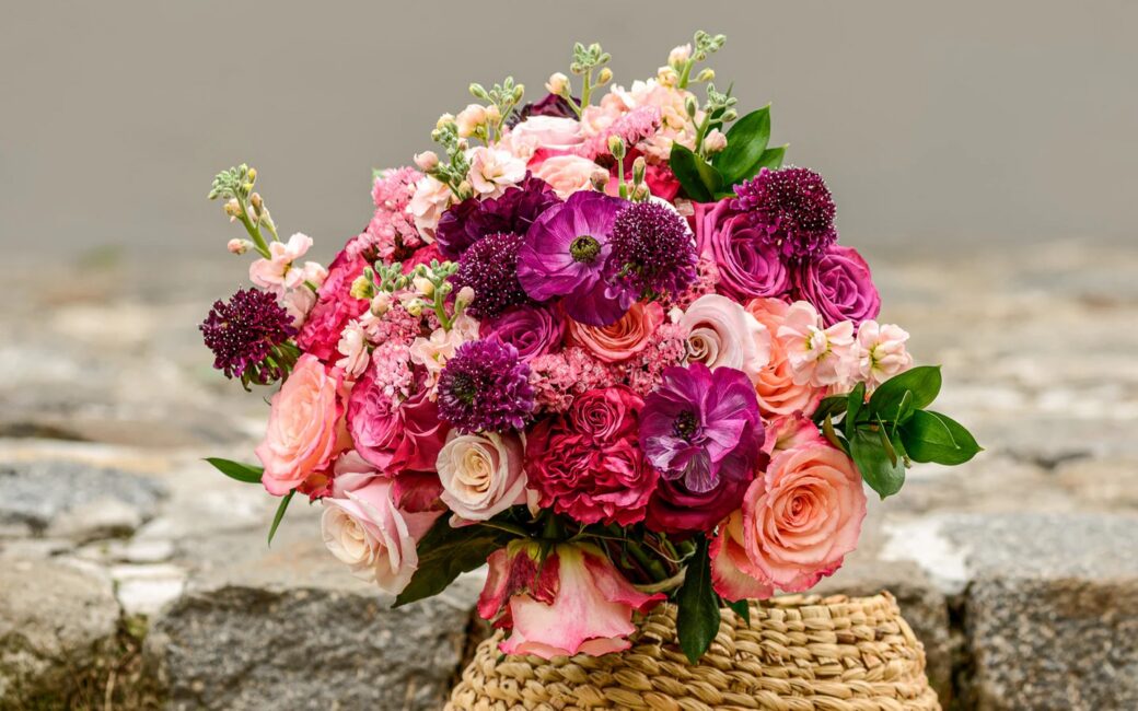 A woven basket holds a bouquet of pink, purple, and cream roses and assorted flowers, displayed outdoors on a stone surface.