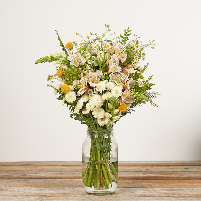 A bouquet of assorted white and yellow flowers in a clear glass jar filled with water, placed on a wooden surface against a plain light background.