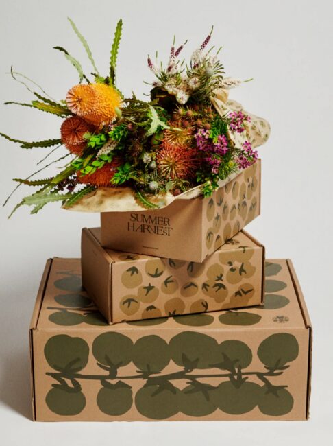 Three stacked cardboard boxes with botanical designs, the top box open and filled with assorted flowers and greenery labeled "SUMMER HARVEST.